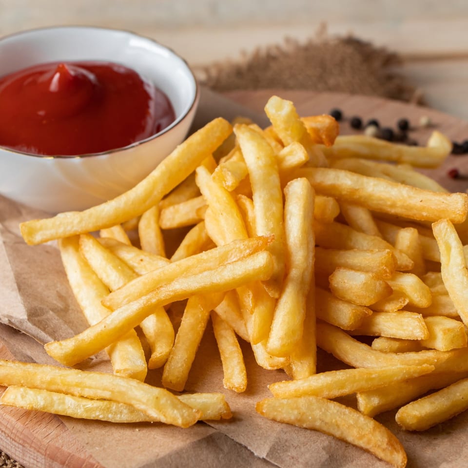 Golden french fries on brown paper with a white bowl of ketchup on a wooden surface.