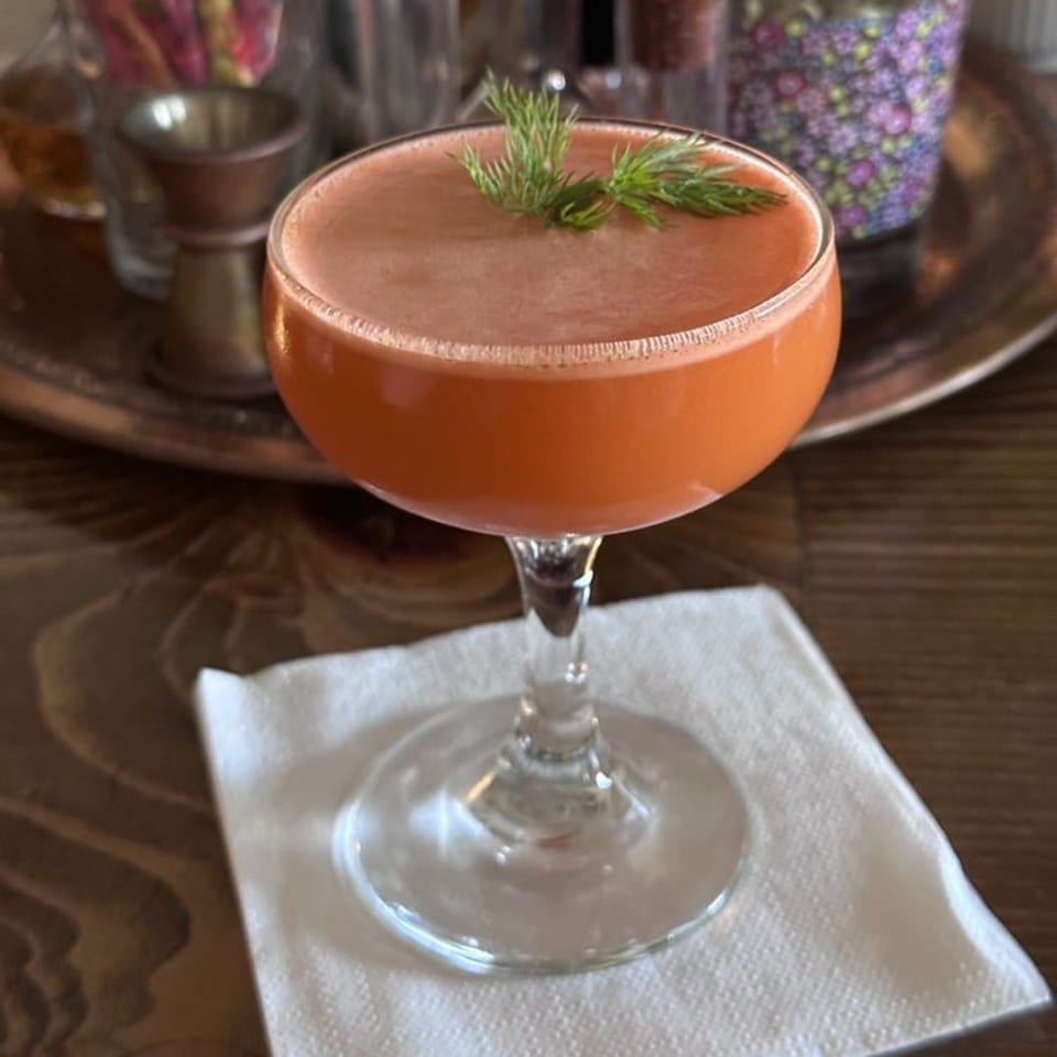 A vibrant orange cocktail, garnished with fresh dill, sits on a white napkin at Lemon Drop.