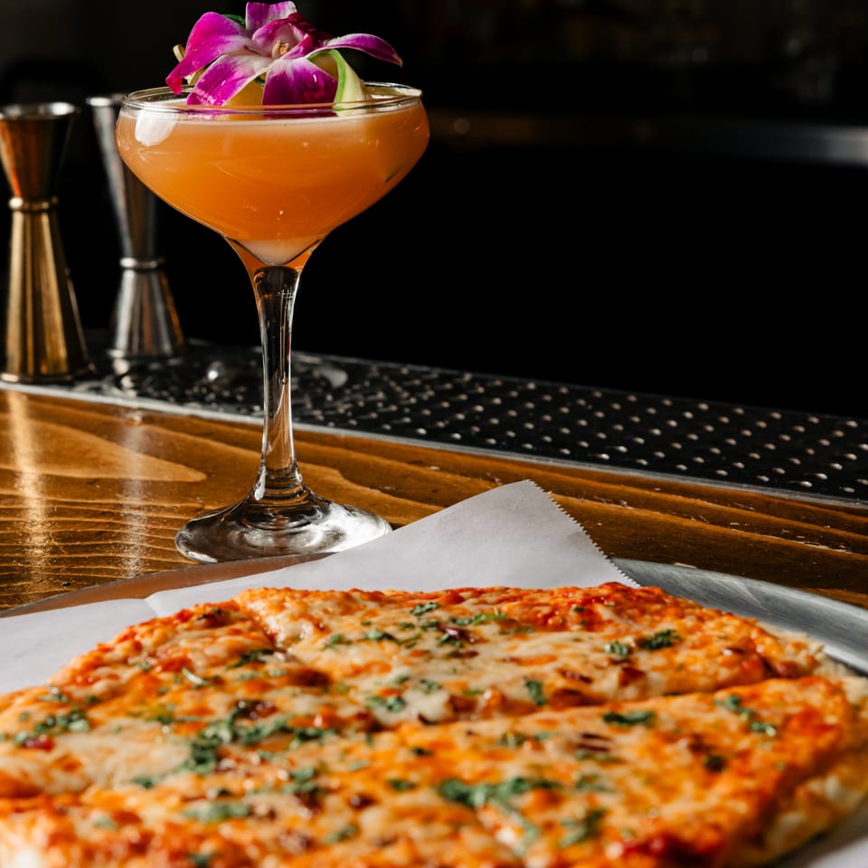 A Lemon Drop cocktail with purple orchids and a flatbread pizza on a wooden bar.