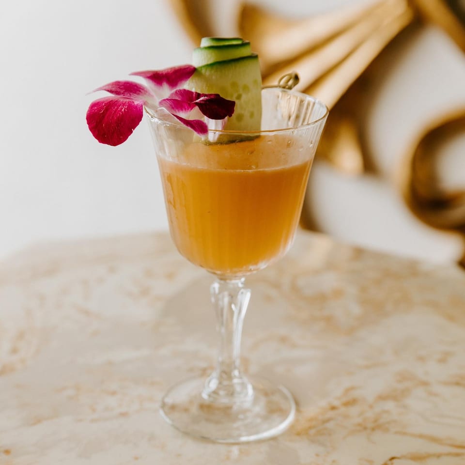 A Lemon Drop cocktail, garnished with a pink orchid and a cucumber ribbon, on a marble surface.