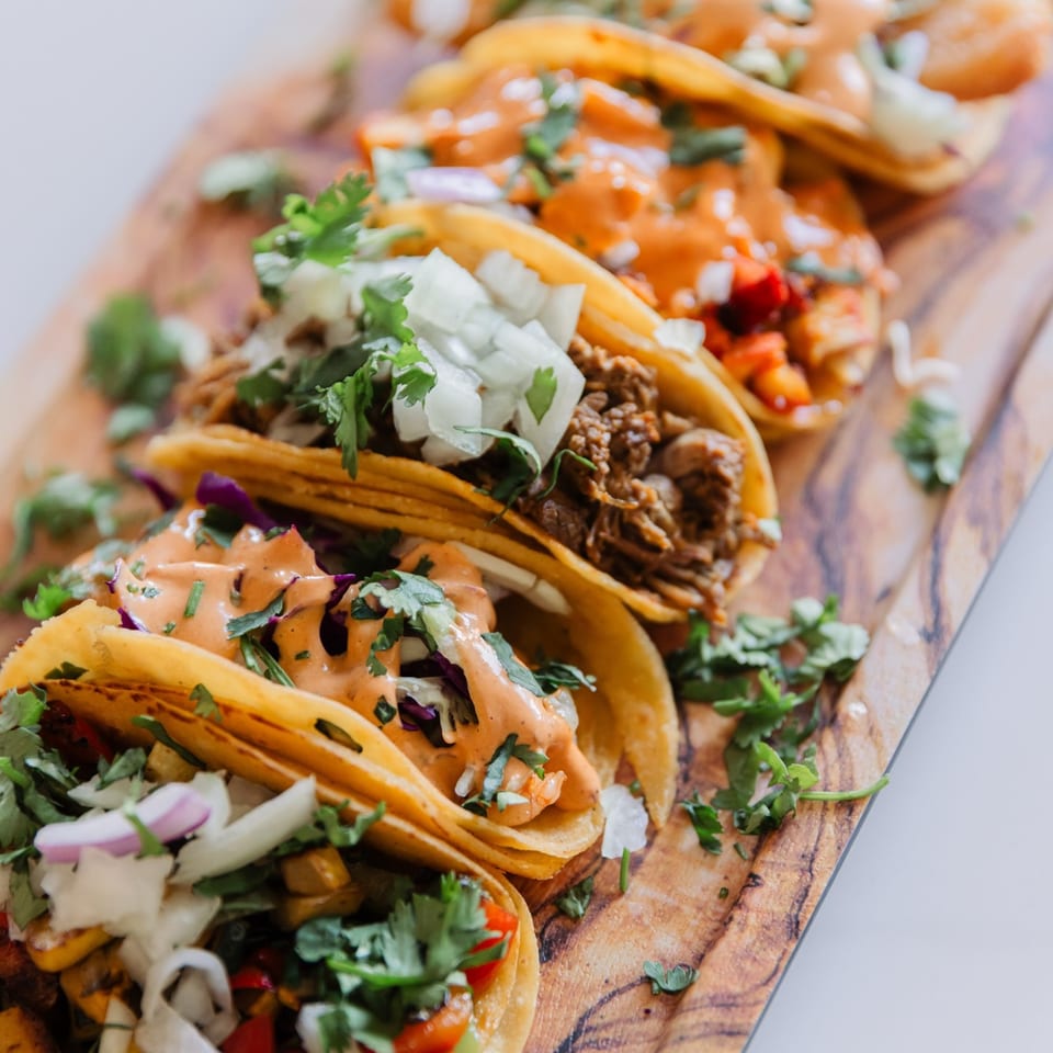A close-up of five vibrant tacos with various fillings, sauces, and fresh cilantro on a wooden board.