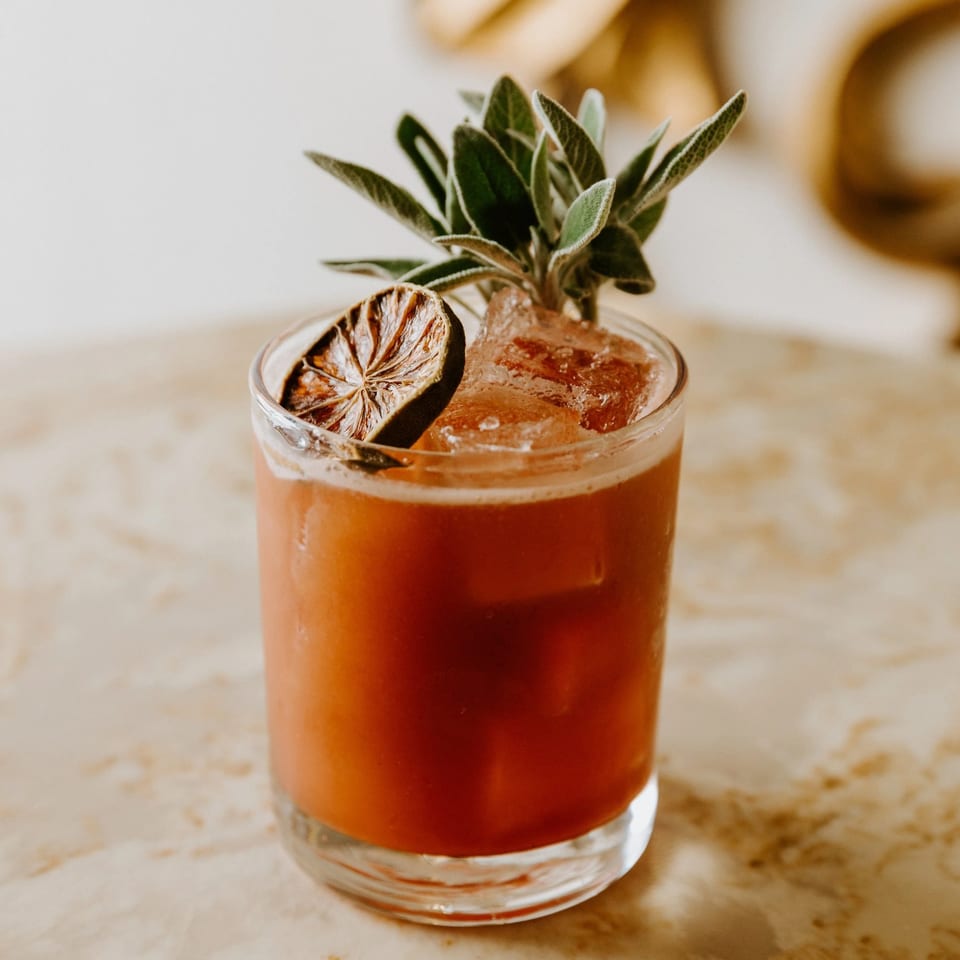 A vibrant red cocktail with ice, garnished with a dried lime slice and fresh sage.