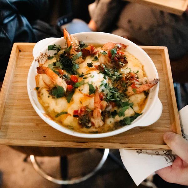A bowl of Lemon Drop's shrimp and grits, garnished with herbs and red peppers, on a wooden tray.
