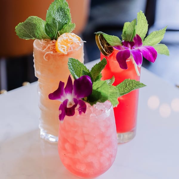 Three colorful cocktails, garnished with mint, citrus, and orchids, on a white table at Lemon Drop.